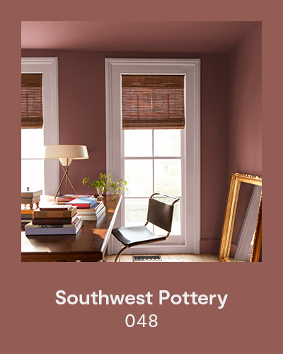 Southwest Pottery 048 - A reddish-brown home office with a wood desk topped with piles of books, a plant, and a lamp with a modern circular lampshade.