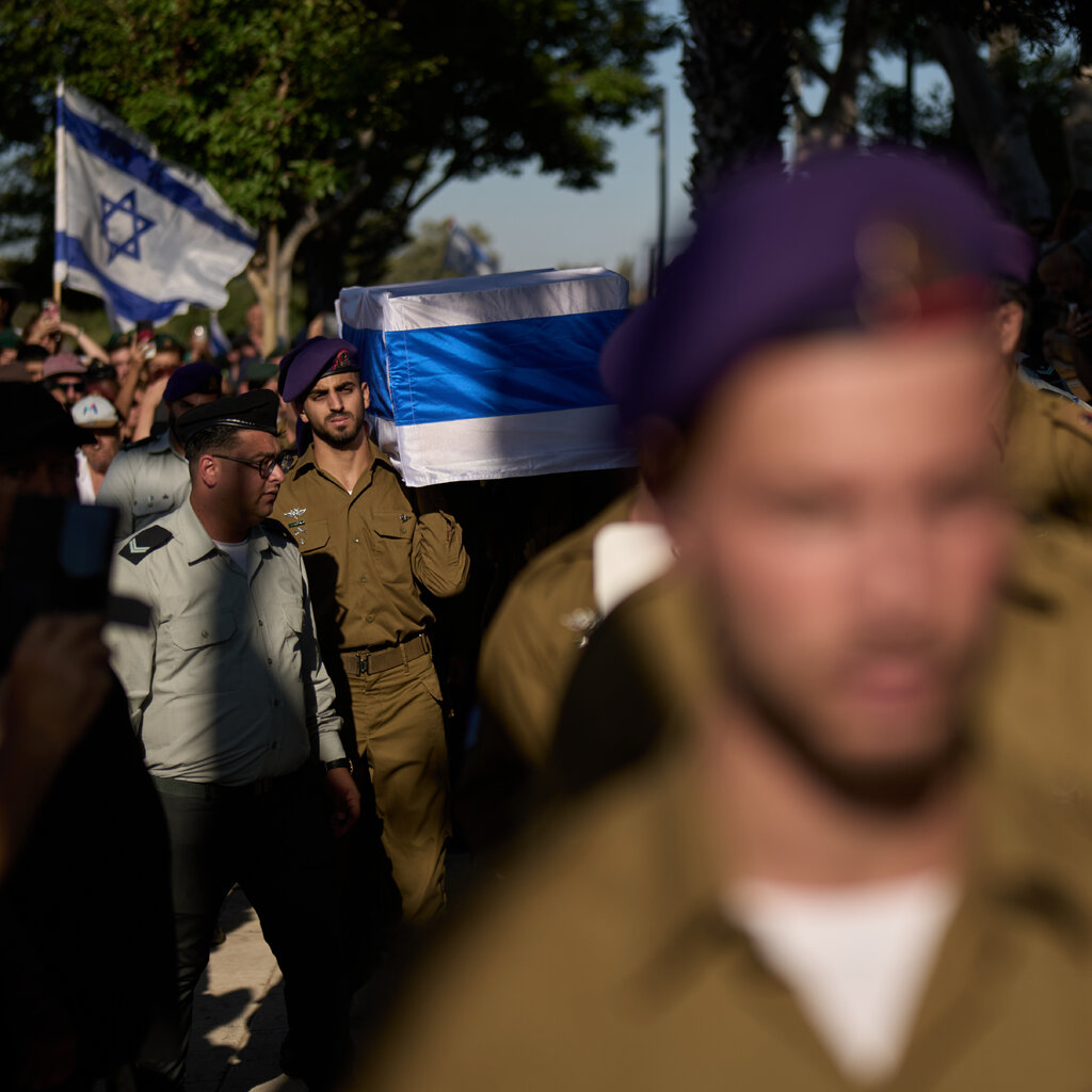 People in uniform with a coffin draped in blue and white pass onlookers. One holds a flag.