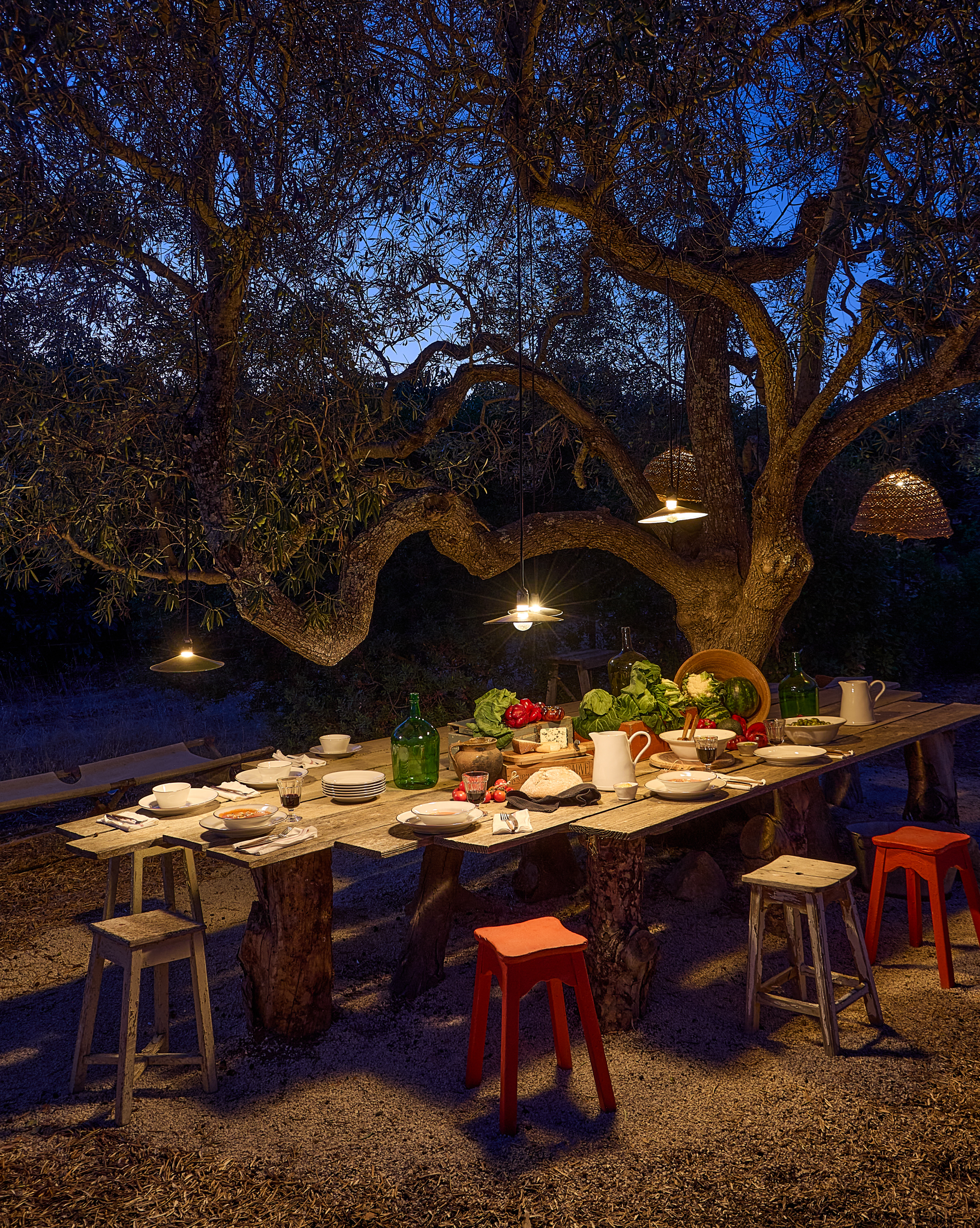 Long outdoor table at dusk, under a tree lit by hanging lamps, with red and white stools, set with Beja tableware, pitchers, green glass demijohns and fresh vegetables