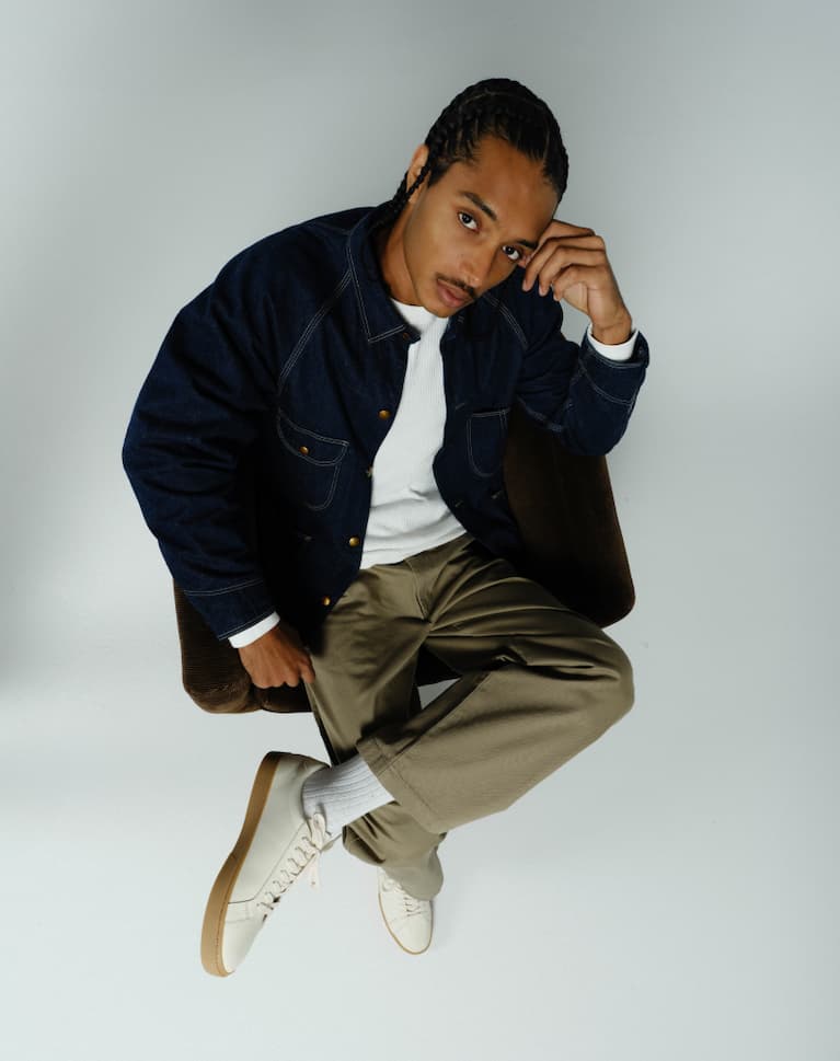 Guy sitting in a chair wearing a dark denim jacket, white tshirt, khakis and white diamond cup sneakers