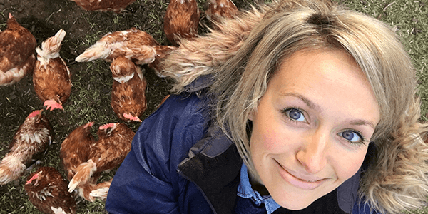 Kate Quilton stands on grass and takes a selfie from above, with lots of chickens walking around her