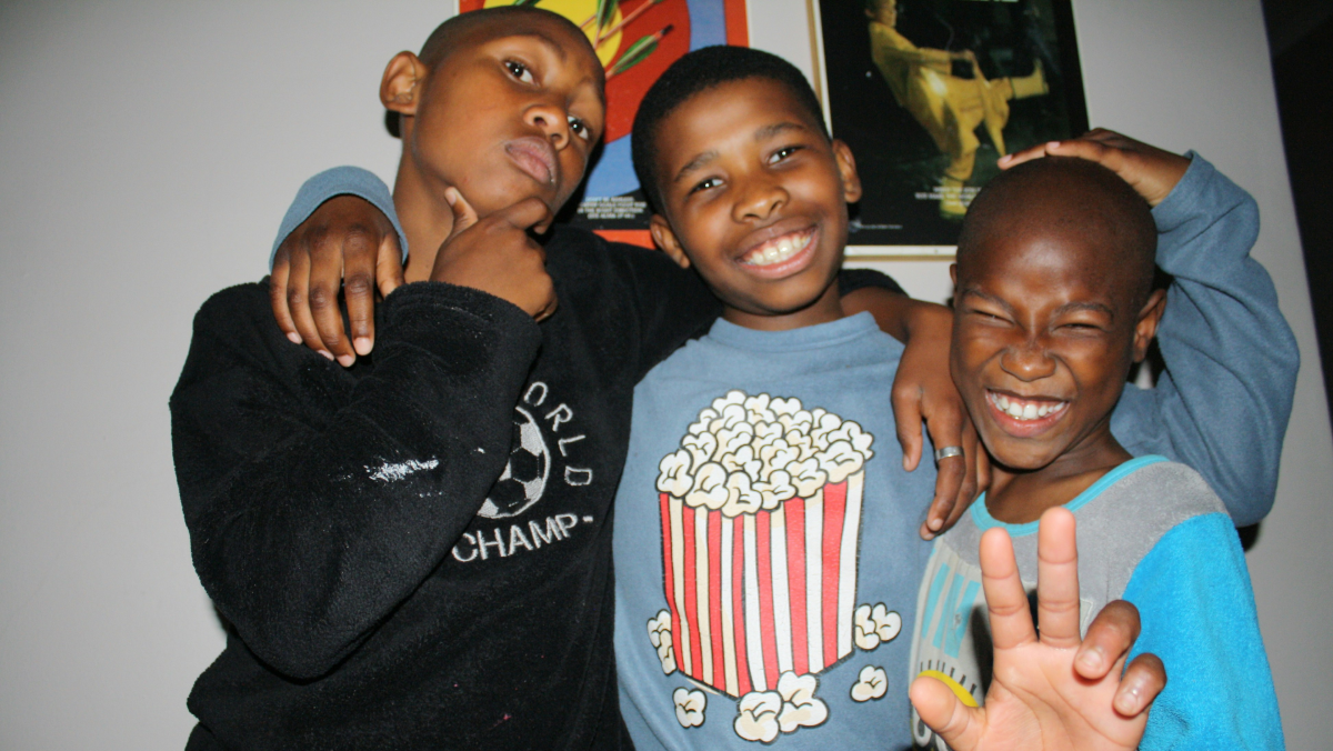 This is a photo of three young boys posing for the camera. They've each got brown skin and close-cropped hair, and the middle boy is putting his arms affectionately around the other two.