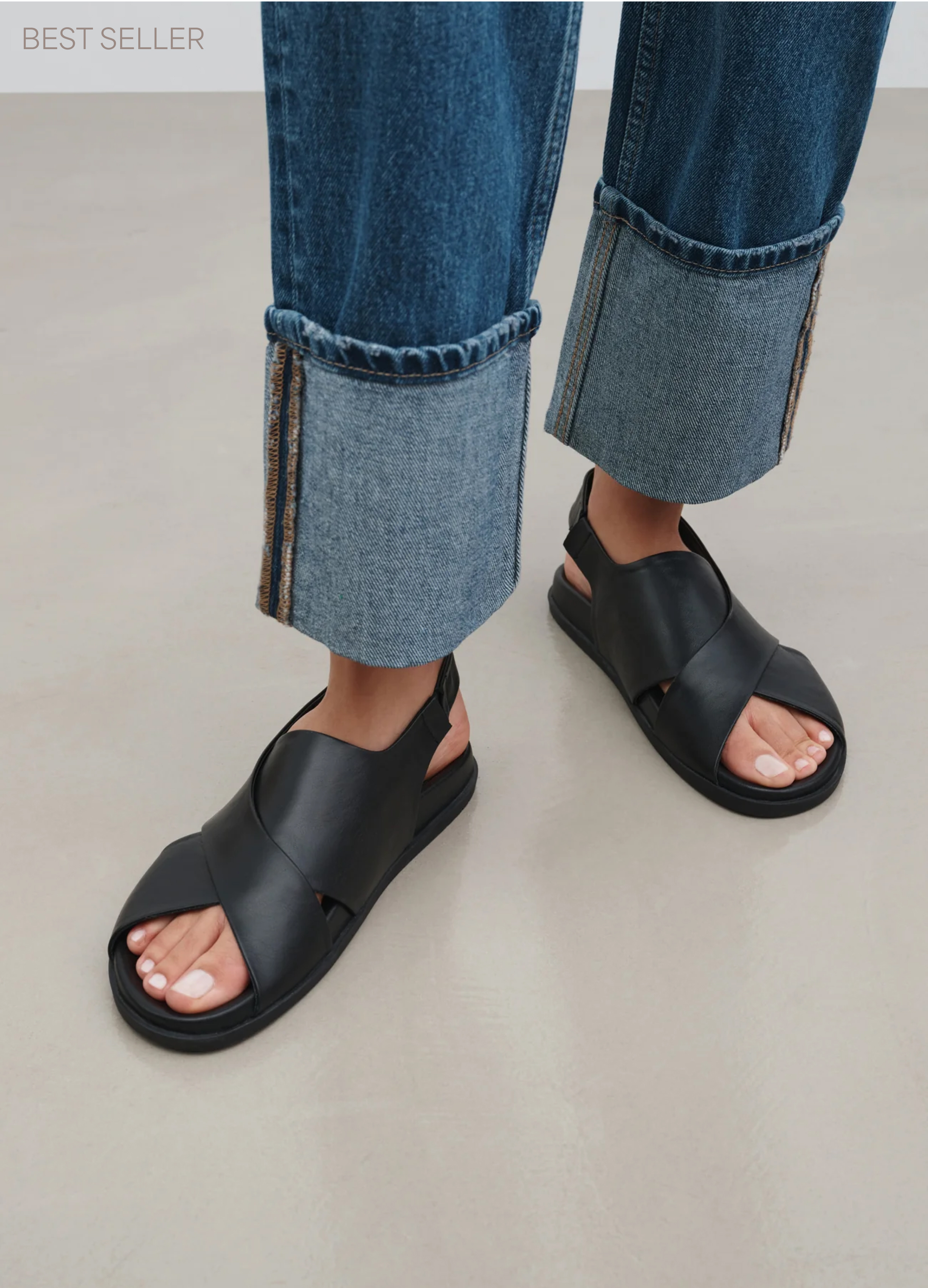 Astrid wears The Heidi Sandals with cuffed jeans, standing on a light gray floor.