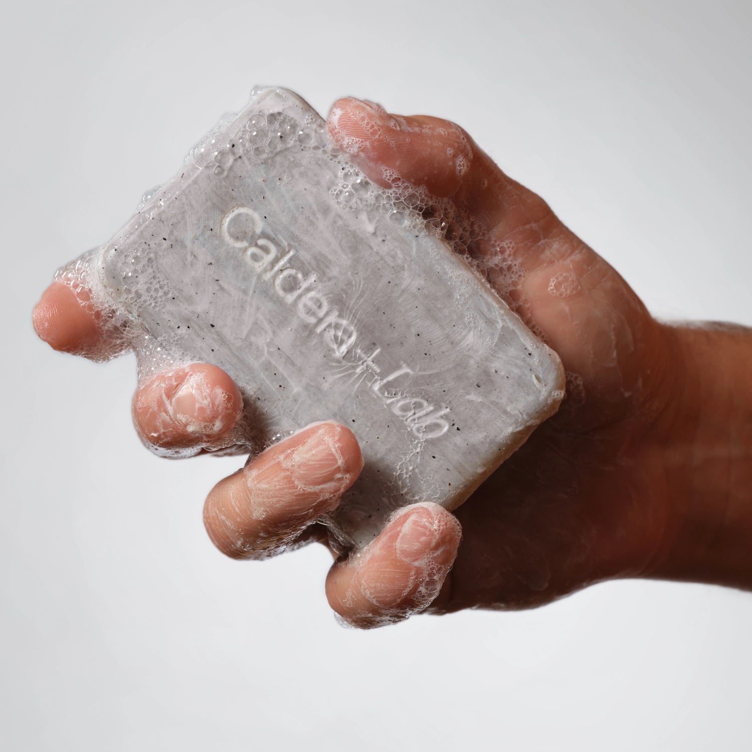 Handle holding a gray colored Body bar by Caldera + Lab, light lather can be seen on the hand and bar and the debossed logo on the bar is pictured.