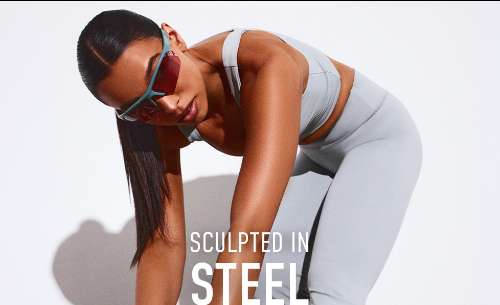 Athletic woman in light grey activewear and teal/red sunglasses, leaning forward. Showcasing new 'Sculpted in Steel' fitness collection.