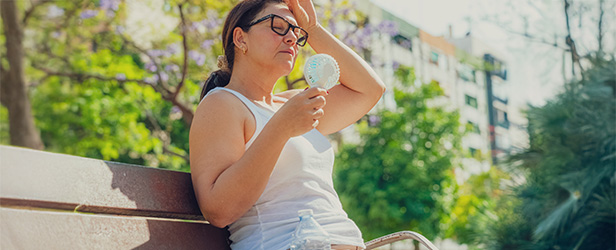 Person sitting on a park bench outdoors, holding a portable fan and wiping their forehead on a sunny day, with trees and buildings in the background.