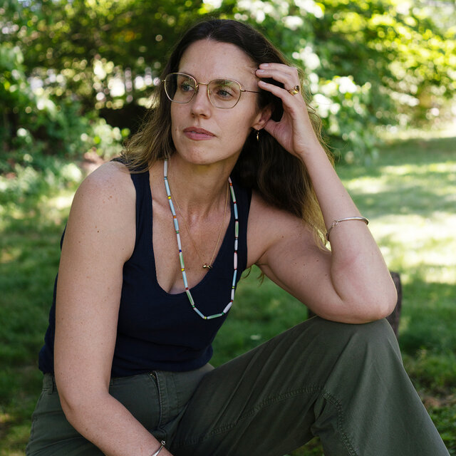 Cybele Malonewears glasses, a black tank top and forest green pants as she kneels on a patch of grass with trees seen in the distance behind her. 