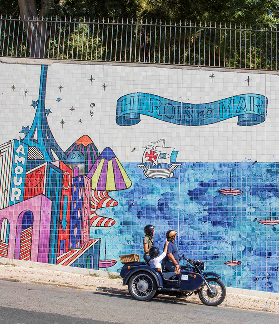 A family riding a motorcycle sidecar passing a vibrant tiled mural along a city street.