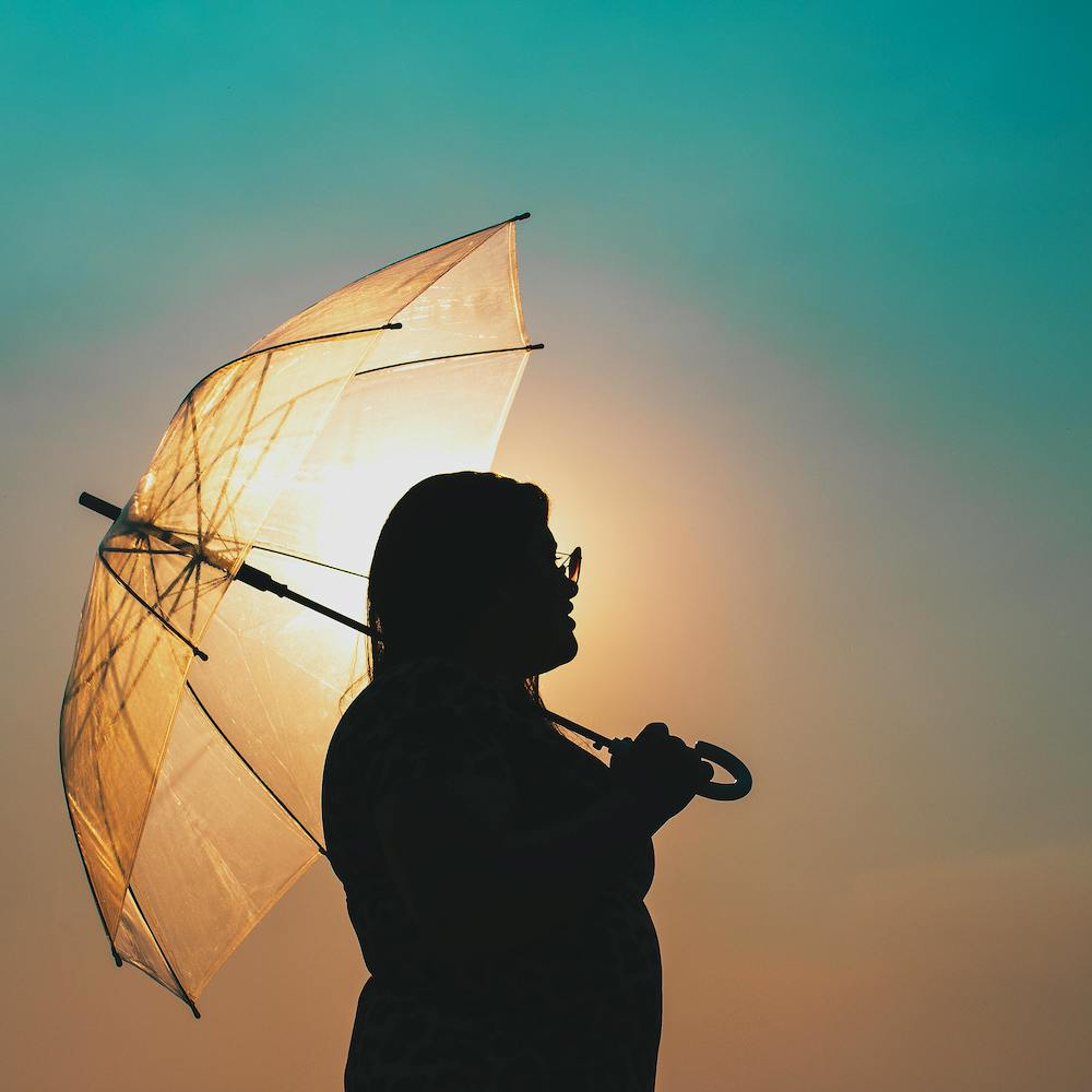 A person silhouetted against the sun holds a clear plastic umbrella open over their head.