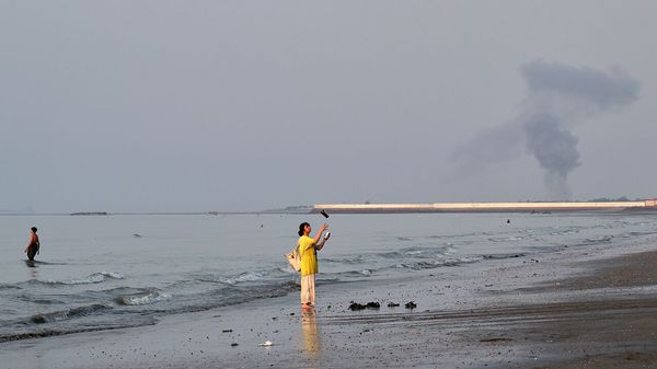 A woman stands at the water's edge along the Strait of Hormuz as a plume of smoke rises in the background following an explosion