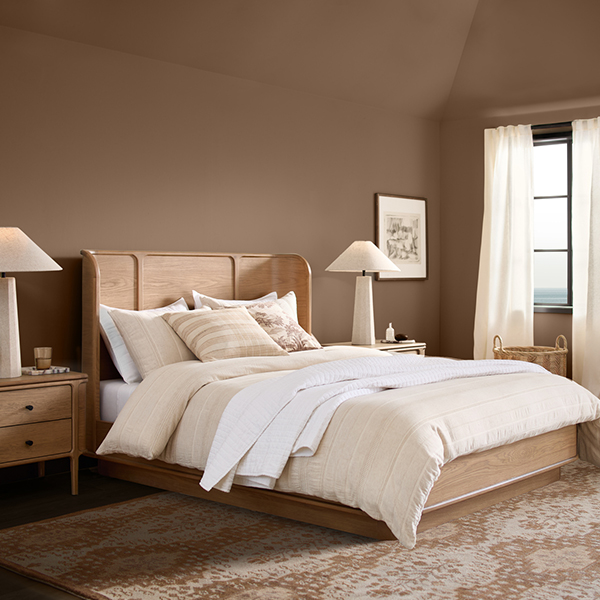 A brown-painted bedroom with a modern platform bed with plush beige linens, matching side tables with lamps, and an area rug.