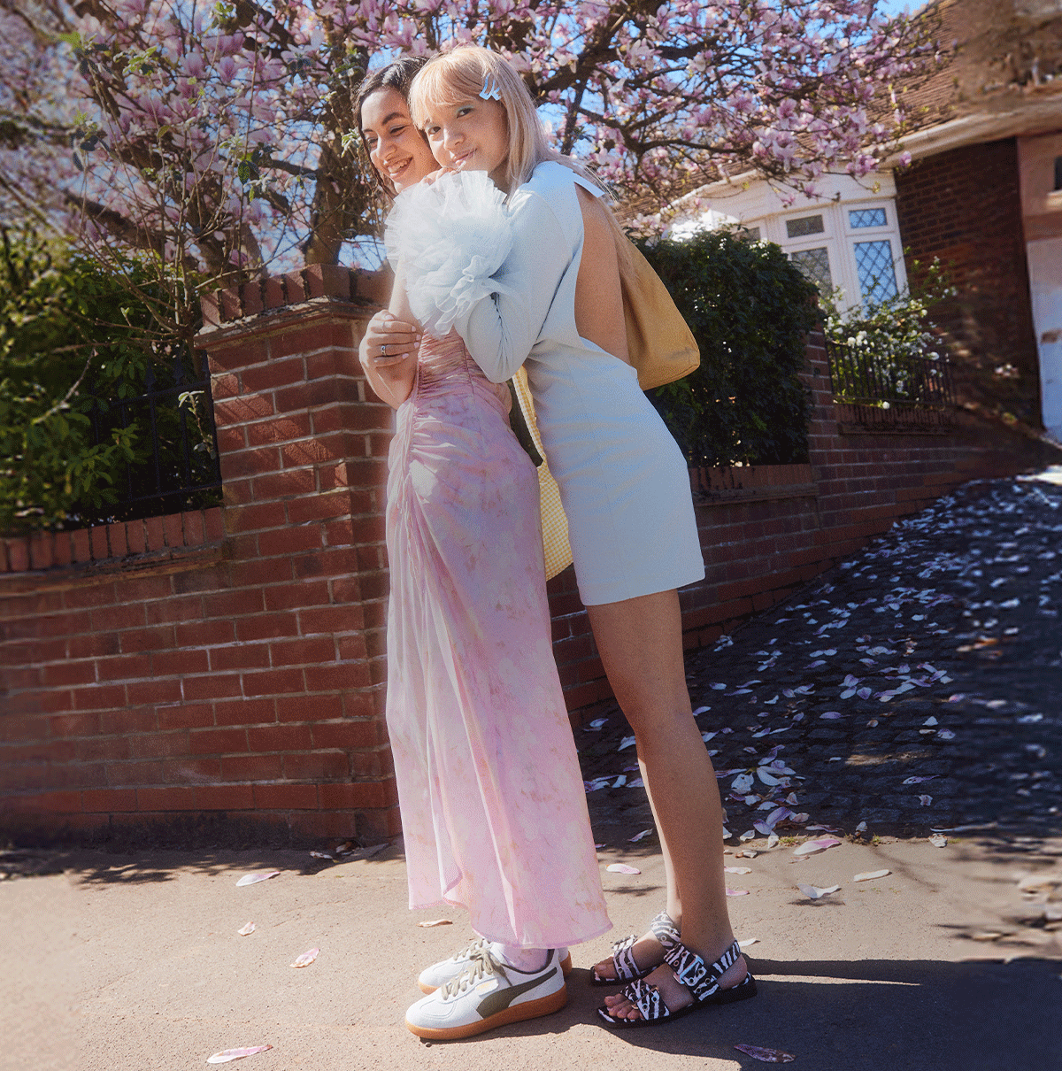 A GIF of two images. First image shows two people wearing pairs of white and black Steve Madden Grand Saria-Le Zebra sandals and light green PUMA Palermo trainers whilst standing in front of a brown brick building. The second image shows the same two people's lower legs wearing the same shoes whilst standing on a concrete ground. The GIF contains the text 'Bank Hol? Treat Yourself'