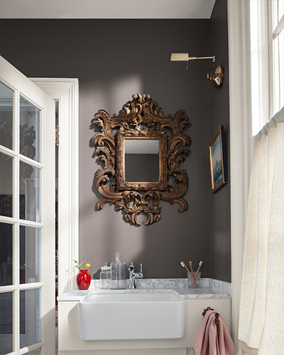 A brown painted powder room with an ornate gold mirror, white vanity, and a window with a gauzy half curtain.