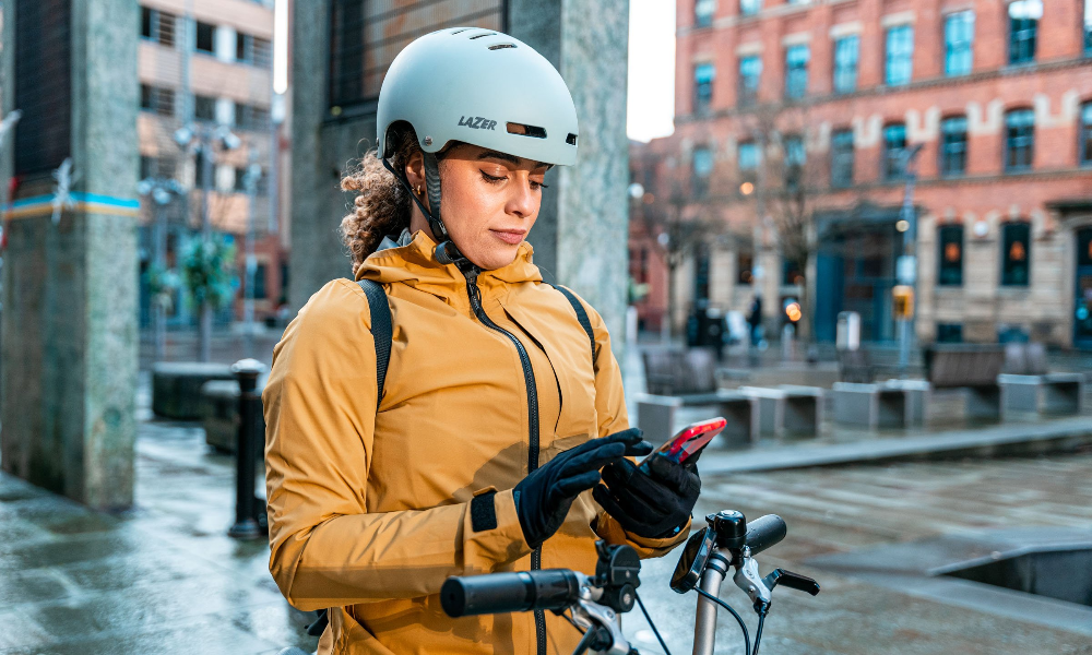 A commuter pauses to look at her phone in a city centre setting, with the handlebars of a folding bike visible in the foreground.