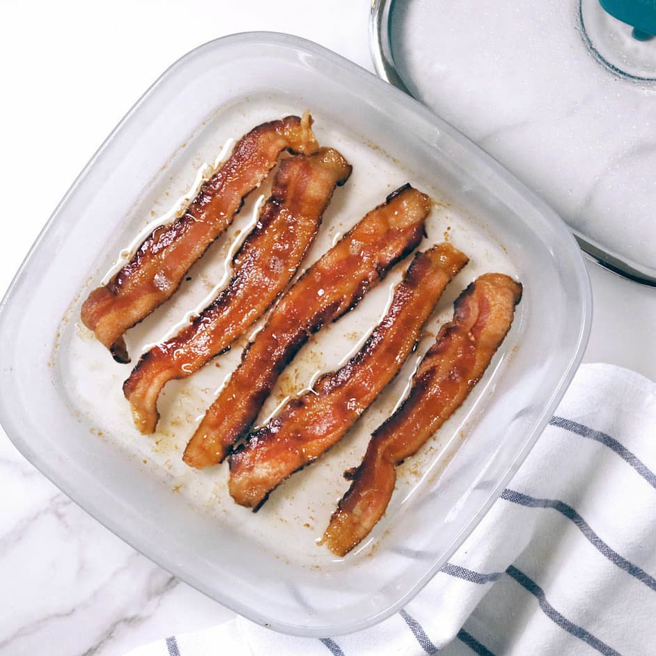  Four crispy strips of cooked bacon sit in an Anyday Square Dish, surrounded by rendered bacon fat. A vented glass lid rests nearby, and a white kitchen towel with navy stripes is partially visible on the marble countertop.