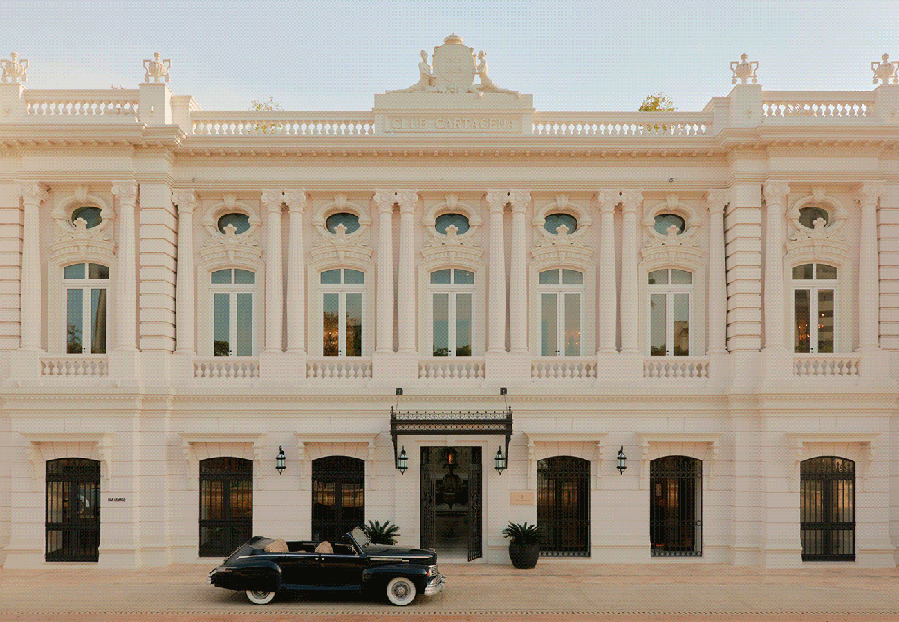 Hero GIF featuring 5 frames: 1) Historic theatre façade with vintage black car parked out front. 2) Tree-lined wooden walkway with person walking between lush greenery. 3) Plated dish with octopus, greens, and sauce on a natural wood table. 4) Man walking down a narrow colonial street lined with colourful balconies. 5) Wide waterfall cascading over cliffs surrounded by dense greenery.
