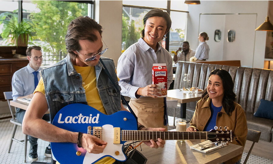 In a diner, a man with a denim vest is playing a Lactaid guitar. The guests and the server smile at him. The server is holding a carton of Lactaid milk.
