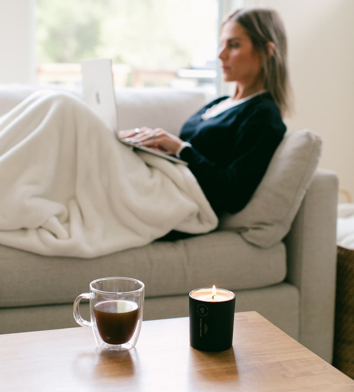 woman sitting on couch on computer with cup of coffee and lit candle on the table in front of her