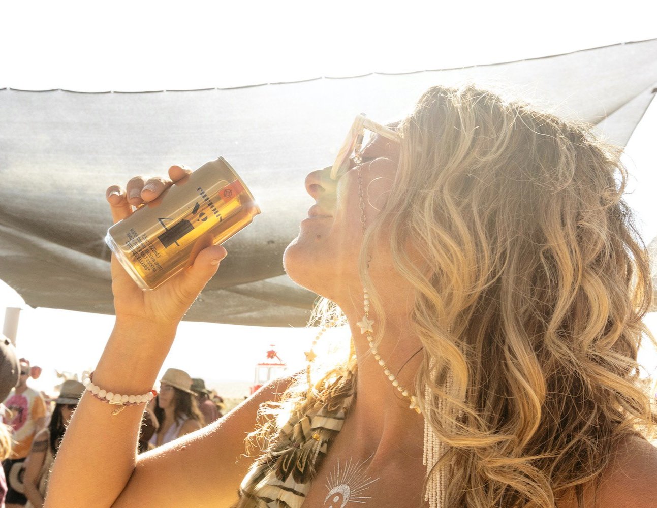 A woman with wavy blonde hair raises a golden mini can of Curious No. 4 toward her mouth as she dances in the sun. 