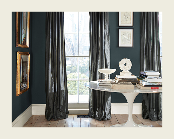 A living space painted dark teal, with similar shaded long curtains and a midcentury modern table topped with books and other accents.
