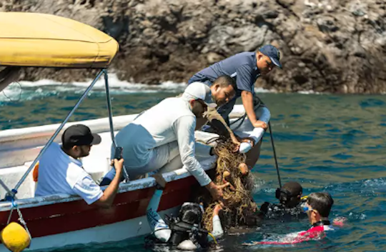 Divers in the water and people on a small boat work together to remove a tangled mass of ghost gear from the ocean near a rocky coastline in Panama