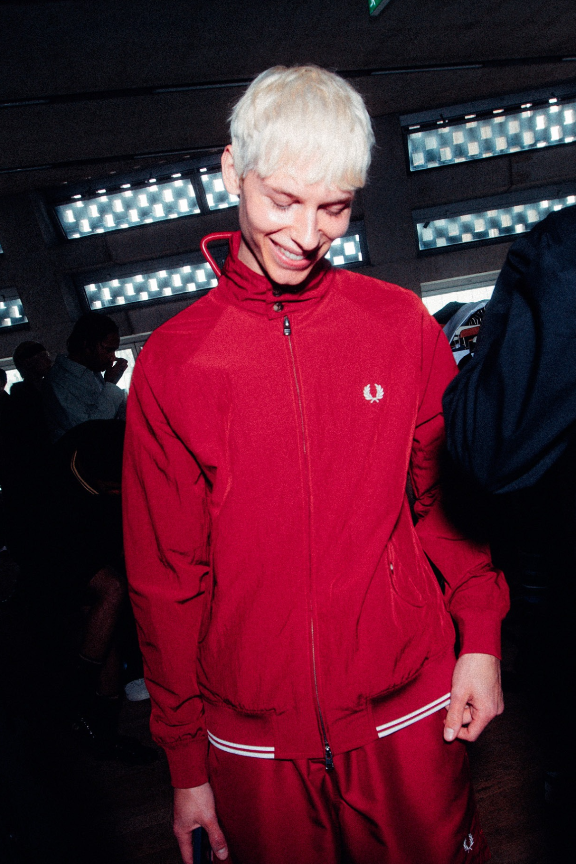 Male white model with short bleached hair wears the red crinkle-textured Harrington Jacket backstage. He is looking down and smiling as the camera flashes.