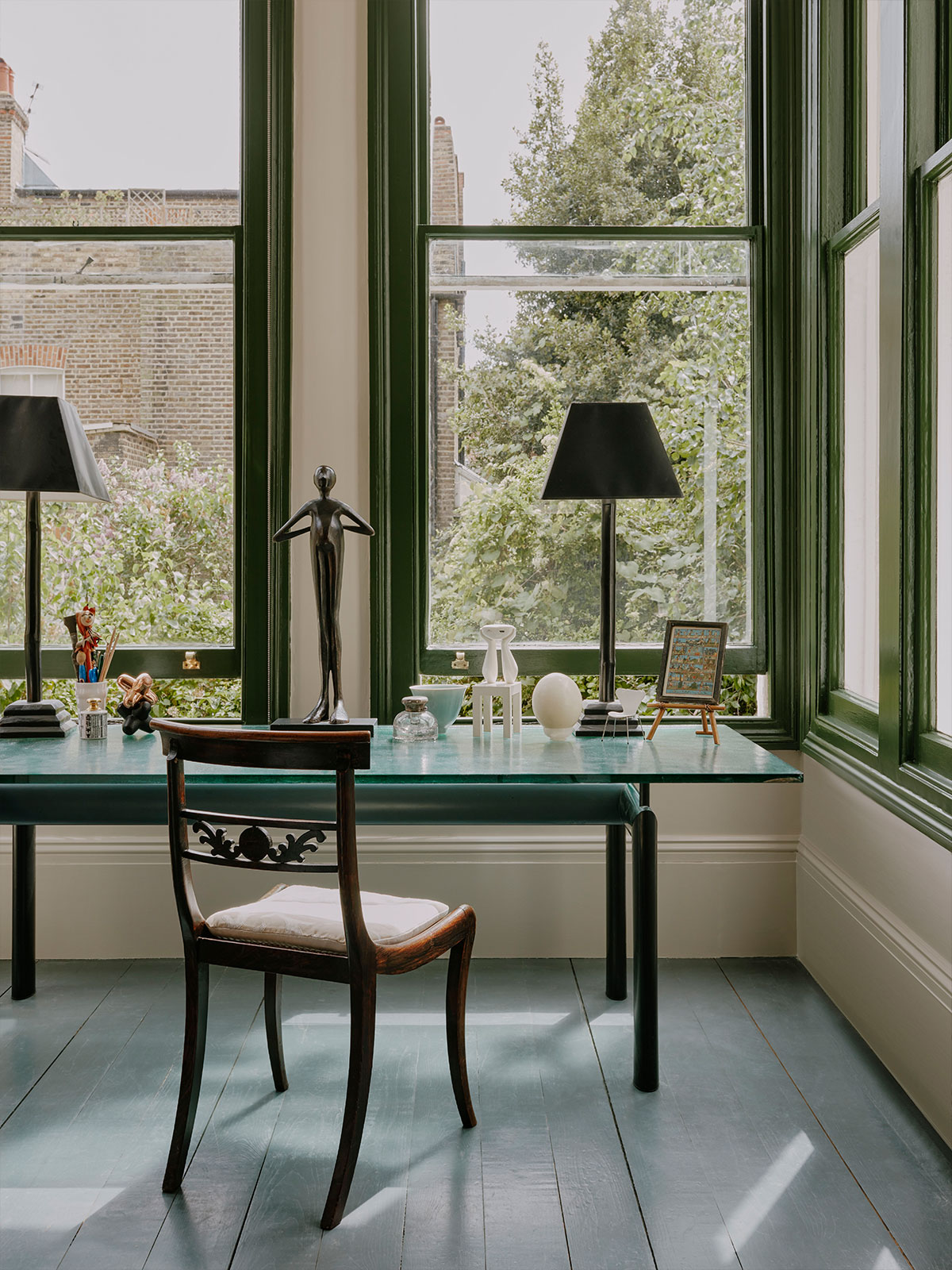 A desk with a view that we might dream of. Light floods in from the double aspect windows, their pea-green wooden frames contrasting with the creamy coloured wall and skirting board. The accessories on the table pick up on the green theme, from the desktop to the lamps, statue, and photograph frame. A serene and inspiring space to work from.