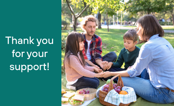 Family with 2 young children holding hands and prayering over a picnic in a park