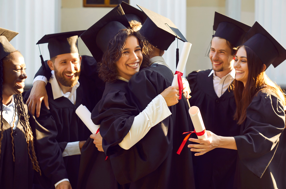 A group of smiling graduates in black caps and gowns celebrate together outdoors, holding rolled diplomas tied with red ribbons.