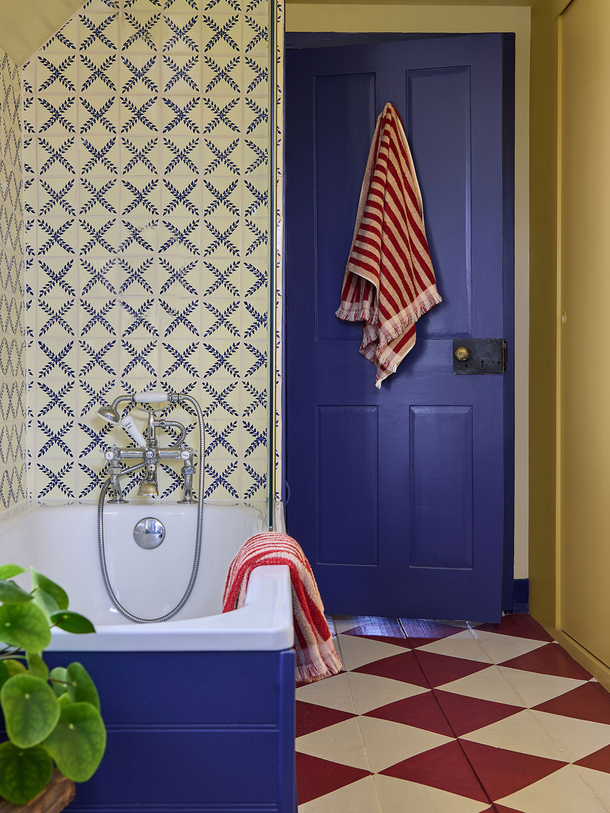 This vibrant family bathroom is a study in contrasting colours and patterns. The vivid blue door matches the wooden panelling around the bath. The chequerboard patterned floor in yellow and crimson contrasts with the pale yellow wall. It’s all offset by fluffy red and white towels ready for wrapping up after bathtime.
