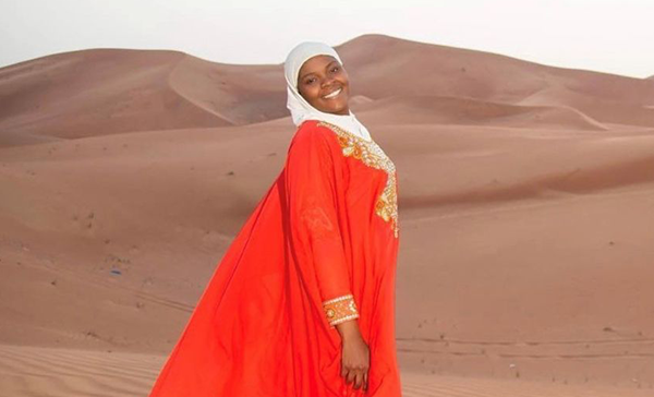 A Black woman poses on a sand dune, wearing an orange dress and white head covering.