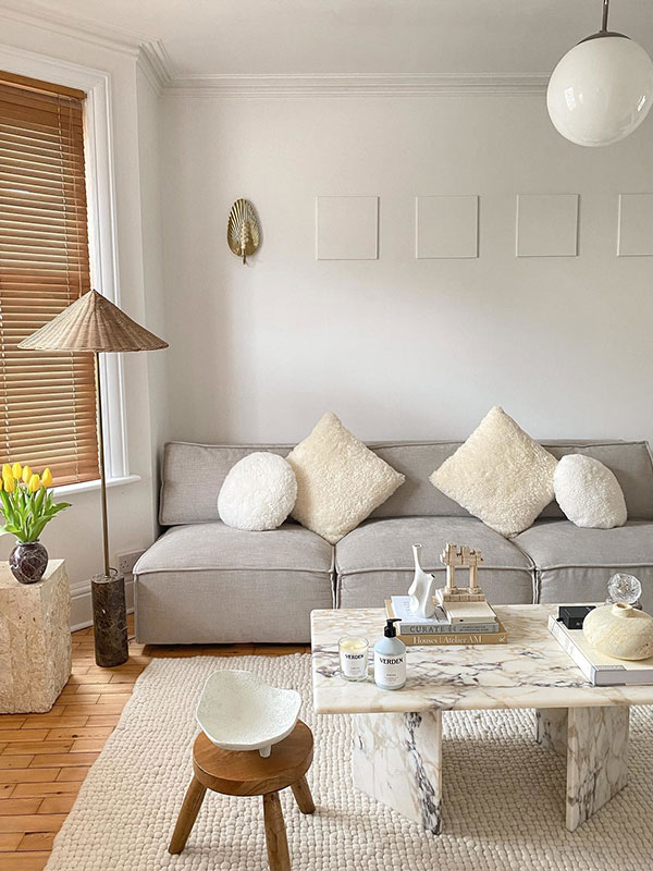 A calm, neutral living room with a light grey sofa, cream textured cushions, and a marble coffee table on a wool rug. Natural wood accents and soft lighting create a warm, minimalist feel.