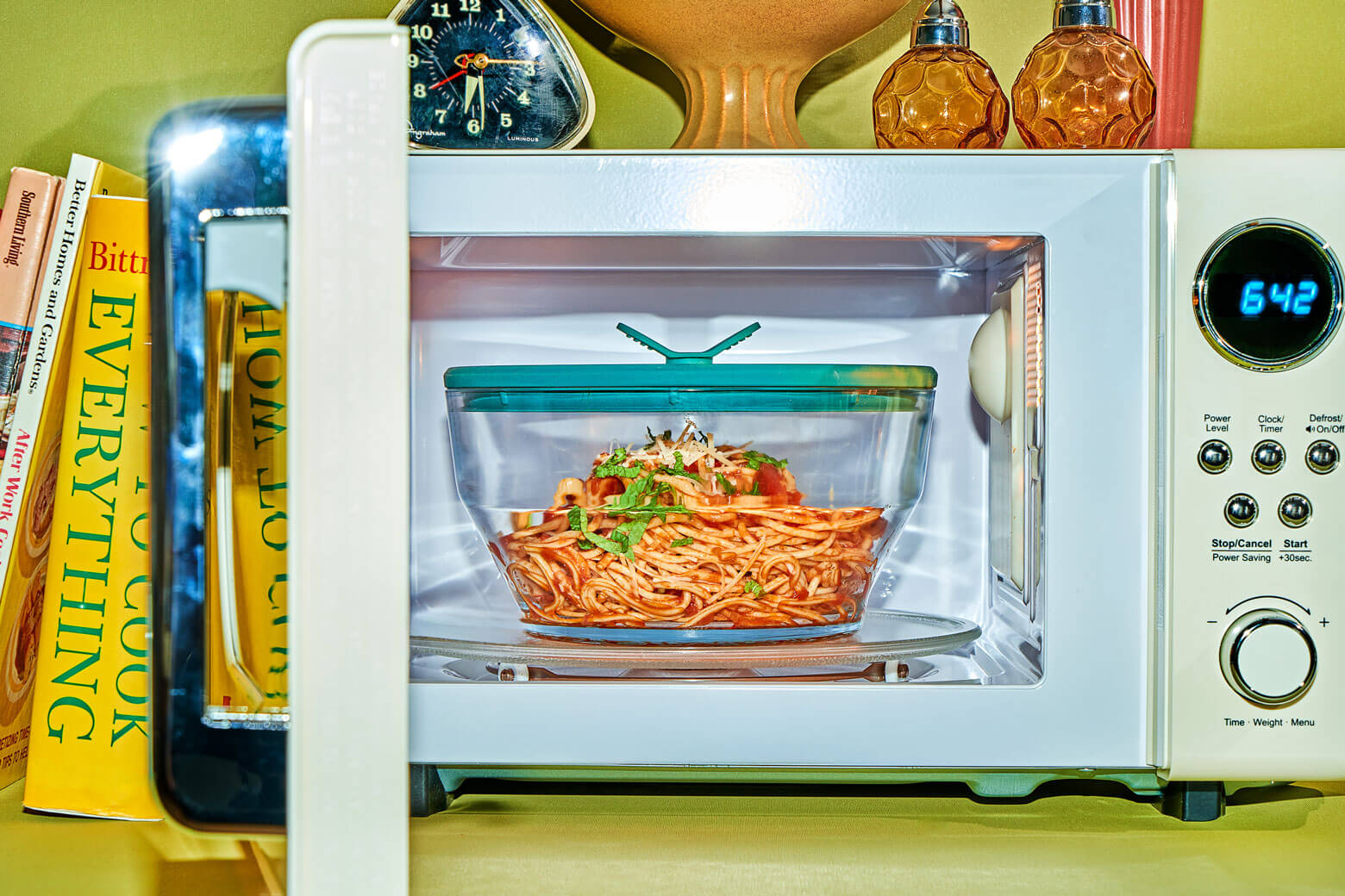 A white microwave with an open door reveals a plastic-free Anyday Everything Dish with a kale-green lid, filled with spaghetti and garnished with fresh herbs. The clear glass container sits on the turntable, showcasing a safe, stain-free solution for microwave cooking.