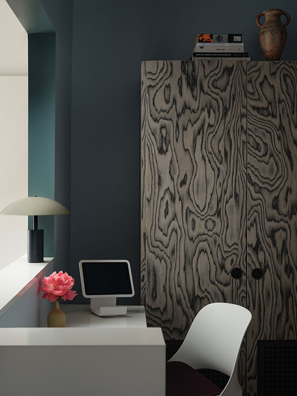 An interior shot of a modern, stylish room featuring a desk setup. On a white desk by a window, a white tablet is on a stand next to a small yellow vase holding a single vibrant pink peony. To the left, a modern black and cream table lamp provides subtle lighting. A large, dark wooden cabinet with a distinctive, swirling wood grain pattern stands to the right. The walls are painted in deep blue and teal tones. The feeling is sophisticated, calm, and conducive to creative work.