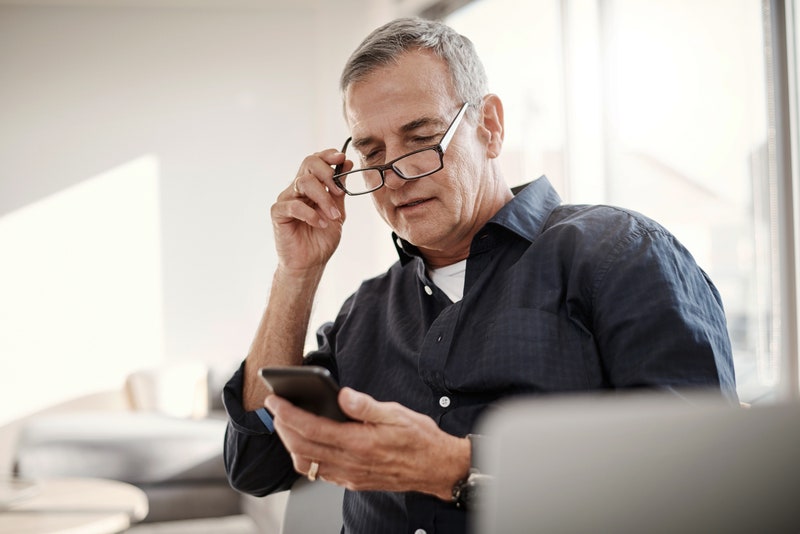 A man takes his glasses off as he looks down at a smartphone. 