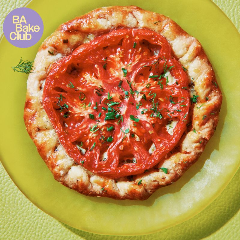 A round of flaky pastry topped with a sliced heirloom tomato, chives, and dill on a plate.