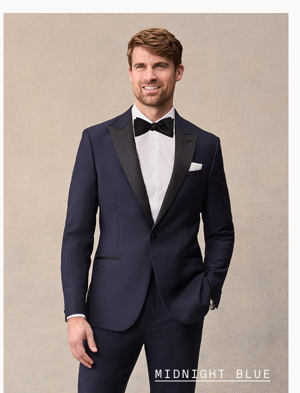 Man with light brown hair smiles, wearing a midnight blue tuxedo with black satin peak lapels, black bow tie, white dress shirt, and white pocket square, hand in pocket. Text overlay reads: "MIDNIGHT BLUE"