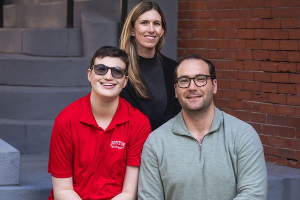 Photo: Three individuals sit and pose for a portrait image. A young man with Down syndrome wearing a red top sits on the left, next to him, SPH Associate Professor of Epidemiology, Eric Rubenstein sits wearing a gray shirt. Behind them, a woman with long hair wearing a black sweater