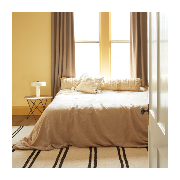 A gold-painted bedroom with a bed on a striped rug in front of a large window with beige curtains, featuring an end table and lamp.