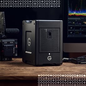 A G-Technology external hard drive, possibly a RAID system, sits on a wooden desk with a computer monitor and camera equipment in the background.