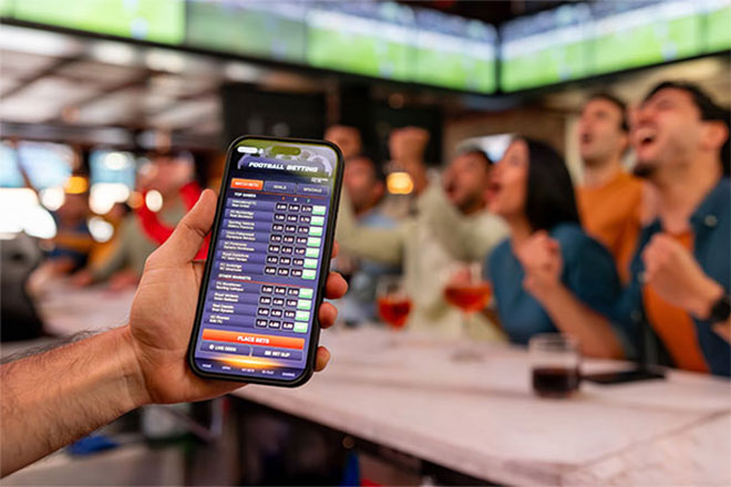 A hand holding a smartphone with a betting app at a crowded sports bar.