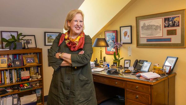 Marsha Guenzler-Stevens in a dark green dress and orange scarf crosses her arms and smiles in front of her desk