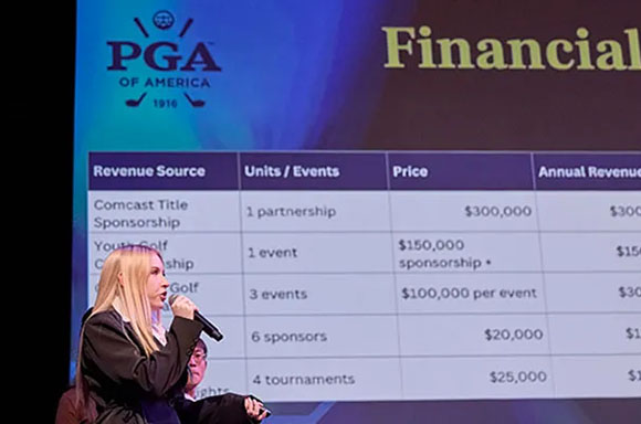 A blonde woman speaks into a microphone on stage in front of a spreadsheet