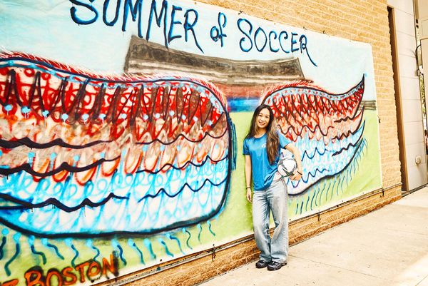 Photo: Carol Gao, a School of Hospitality Administration student and an intern at Absolut Sports, posing in front of a wall that says "Summer of Soccer" with colorful angel wings