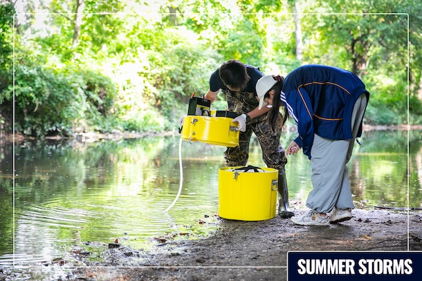 Two students on a sunny day at Cobbs Creek using research equipment. "Summer Storms" text is in the lower-right corner and the image is surrounded by a light rectangular frame.