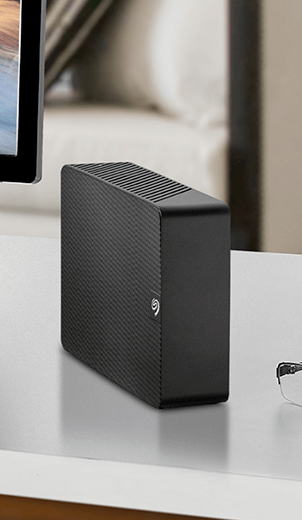 A Seagate Expansion Desktop sits on a desk beside a computer monitor and some glasses.