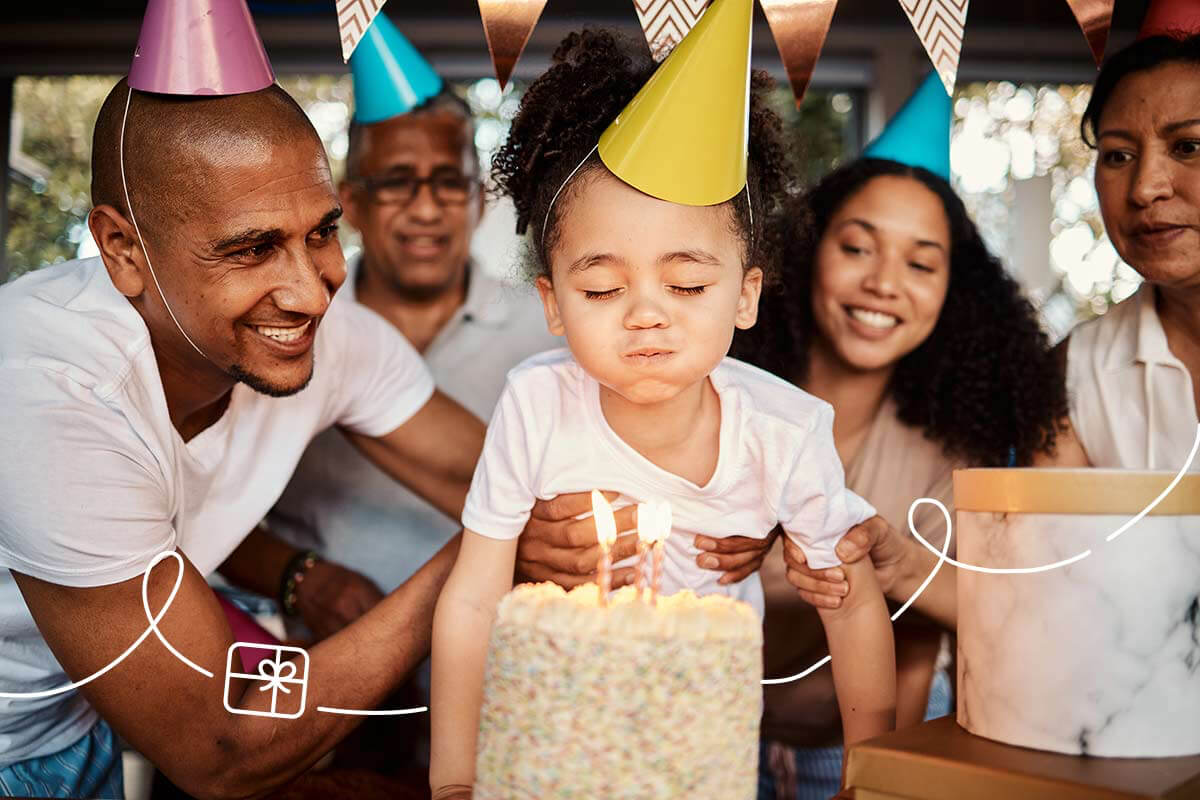 A child blows out candles on a cake while a group of partygoers smile and watch attentively.