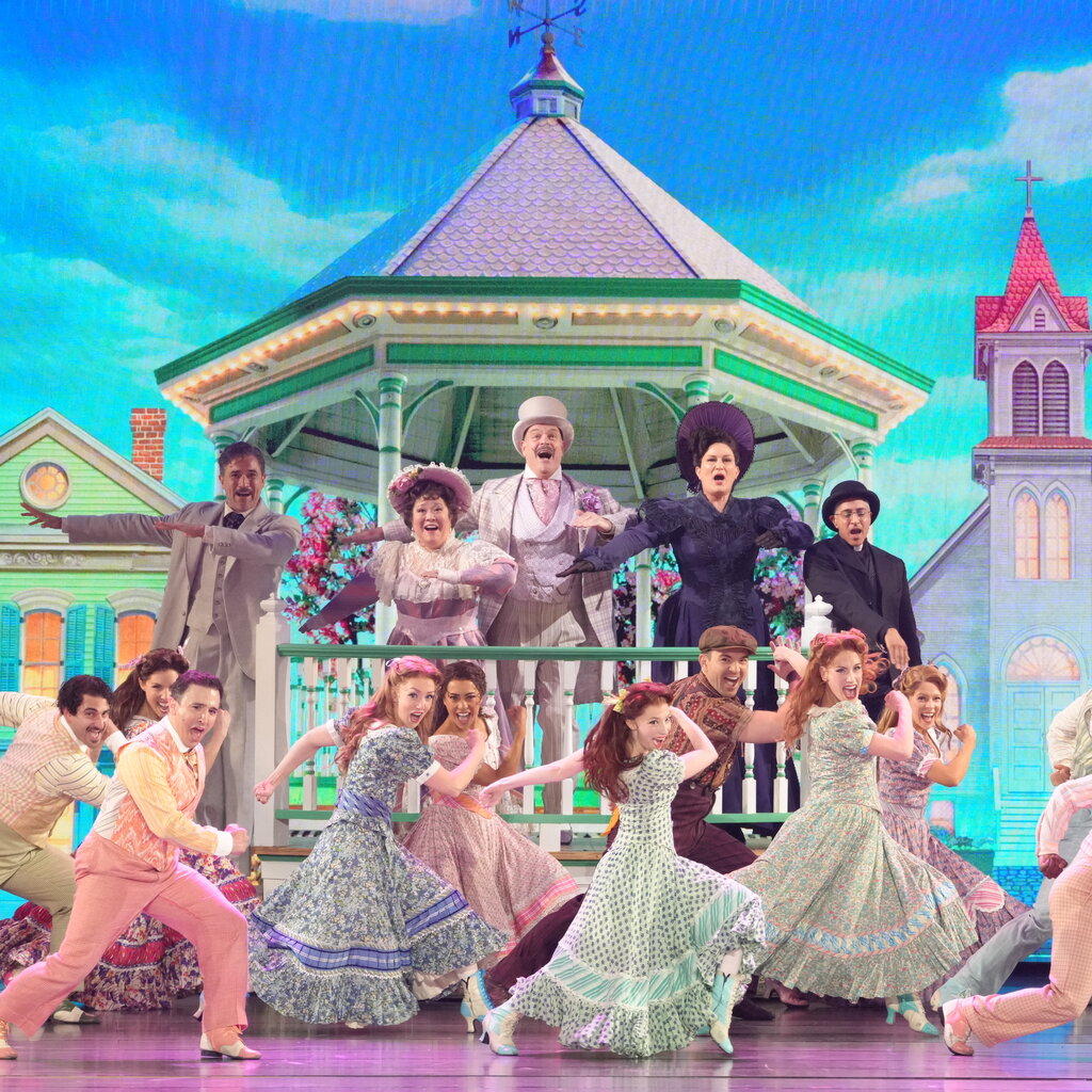 A group of people in brightly colored period costume sing and dance on a stage.