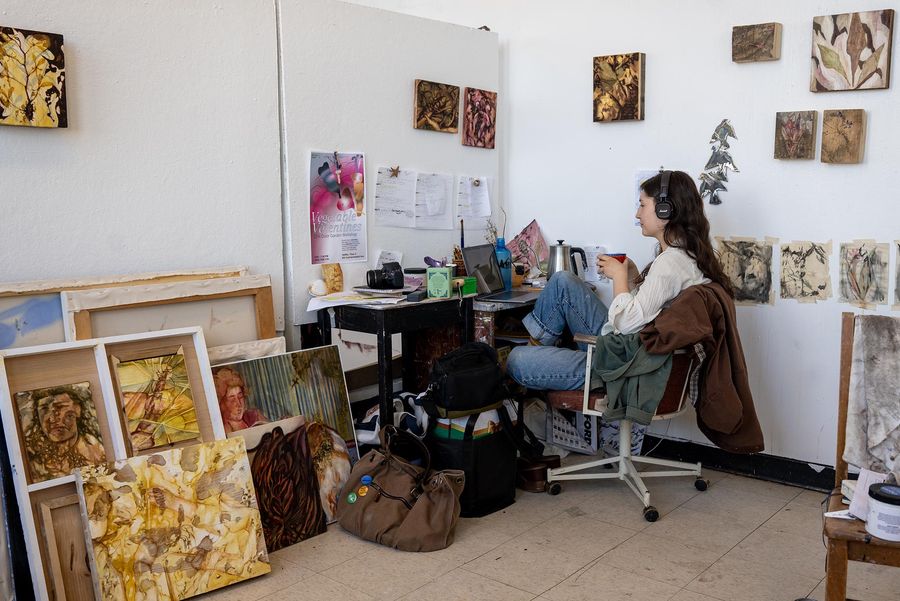 Photo: A woman sits at her desk wearing headphones while holding a red mug. She is surrounded by her art on the walls and organized on the floor