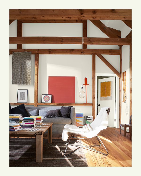 Exterior light streams into an inviting living room painted in white hues, exposed wood beams, gray couch, wood floor, and pops of color throughout.